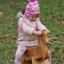 Téli sapkát és kabátot viselő kislány lovagol egy egyszerű, natúr fából készült hintalovon a füvön.